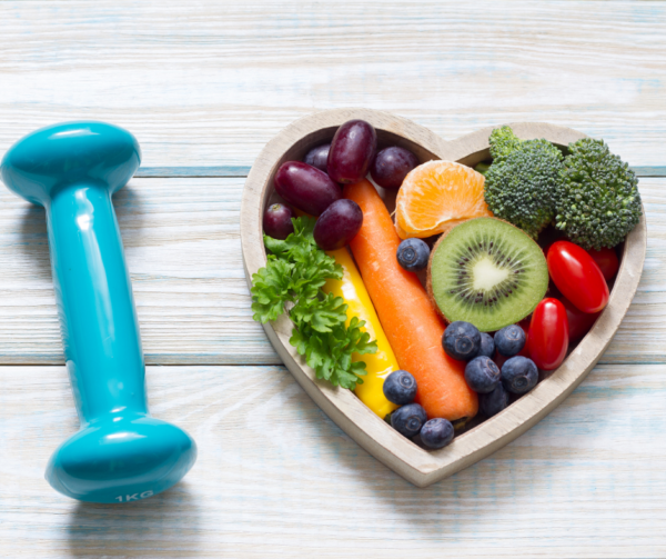 Heart Shaped wooden bowl full of fresh fruit and vegetables with a dumbell resting next to it